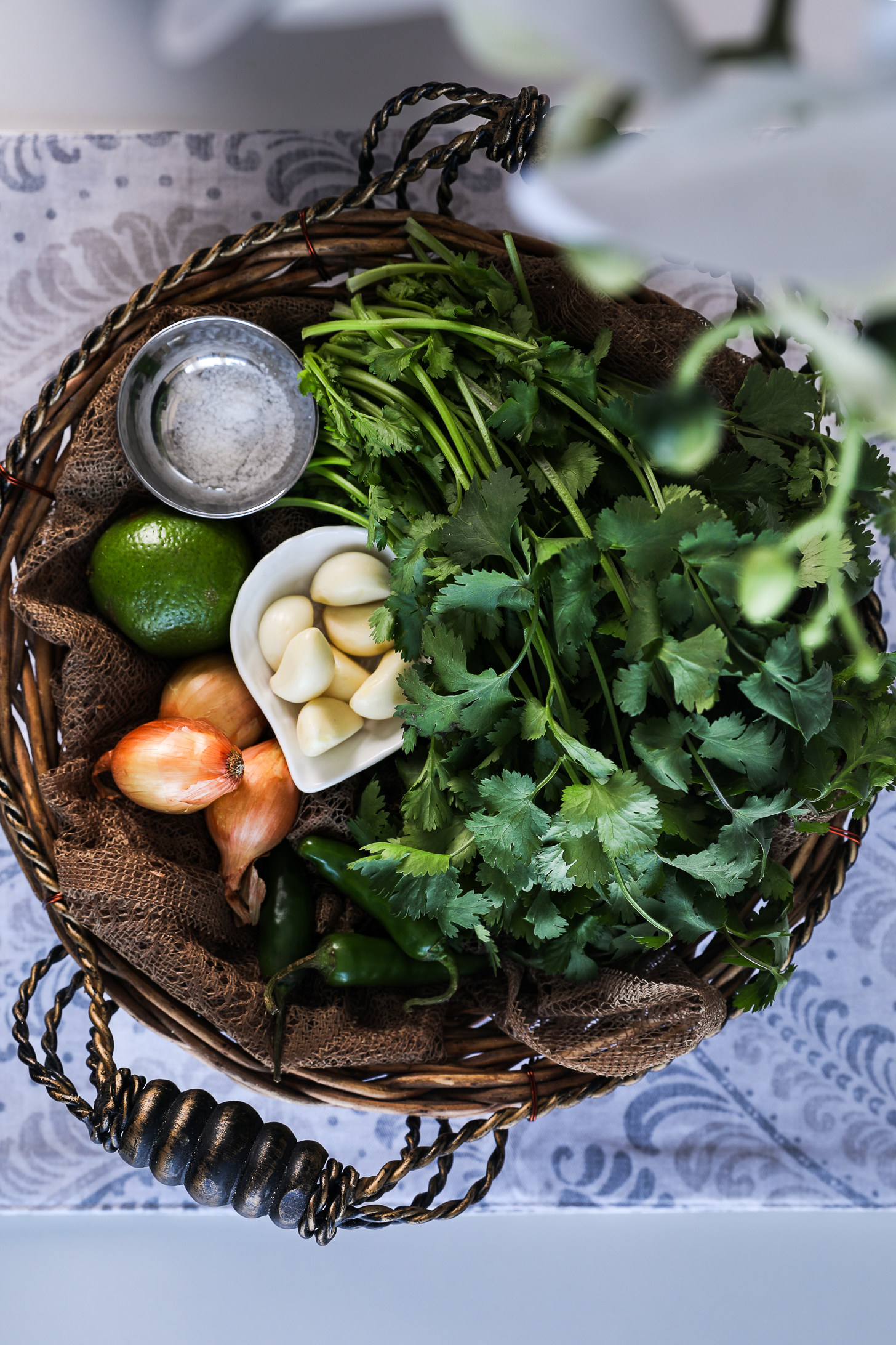 Top view image of a basket of food ingredients including fresh cilantro, garlic, shallots, lime, chillies and salt on a white printed tablecloth.