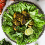 Overhead close-up image of fried green chicken breast on lettuce, adorned with chillies and lime, styled with an Indian scarf and cilantro.