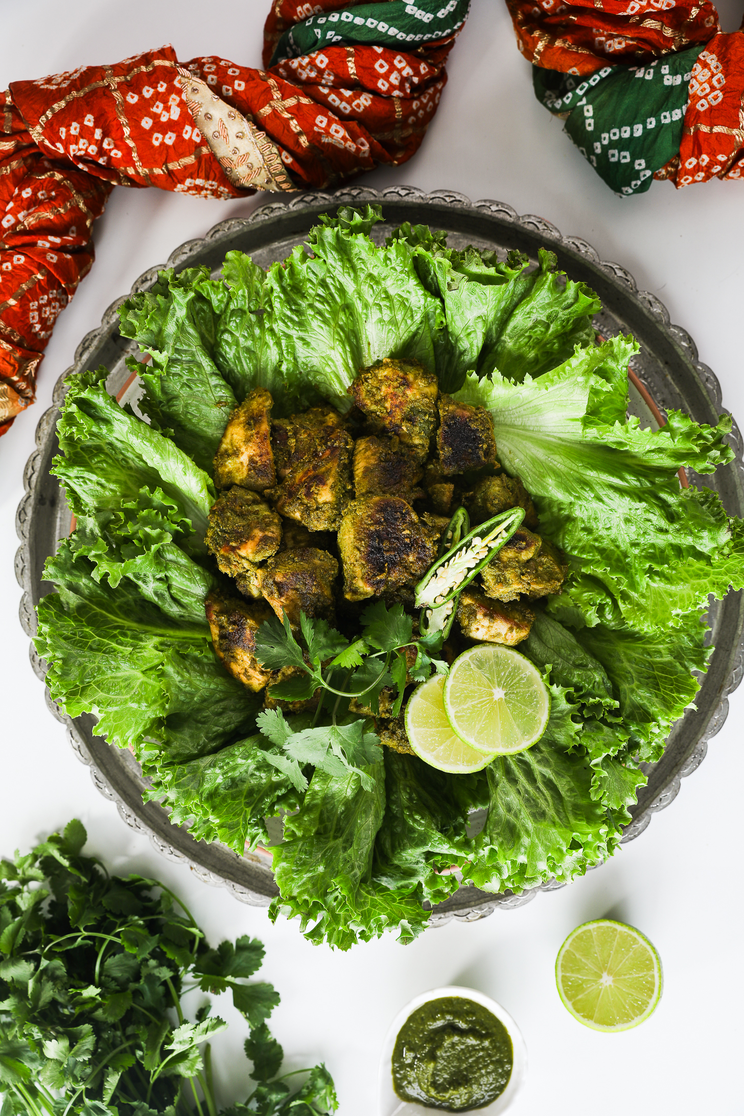 Overhead close-up image of fried green chicken breast on lettuce, adorned with chillies and lime, styled with an Indian scarf and cilantro.