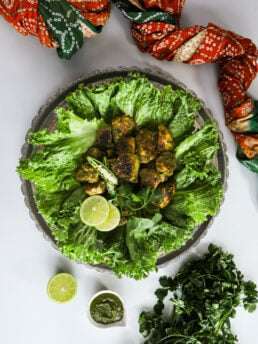 Overhead image of fried green chicken breast on lettuce, adorned with chillies and lime, styled with an Indian scarf and cilantro.