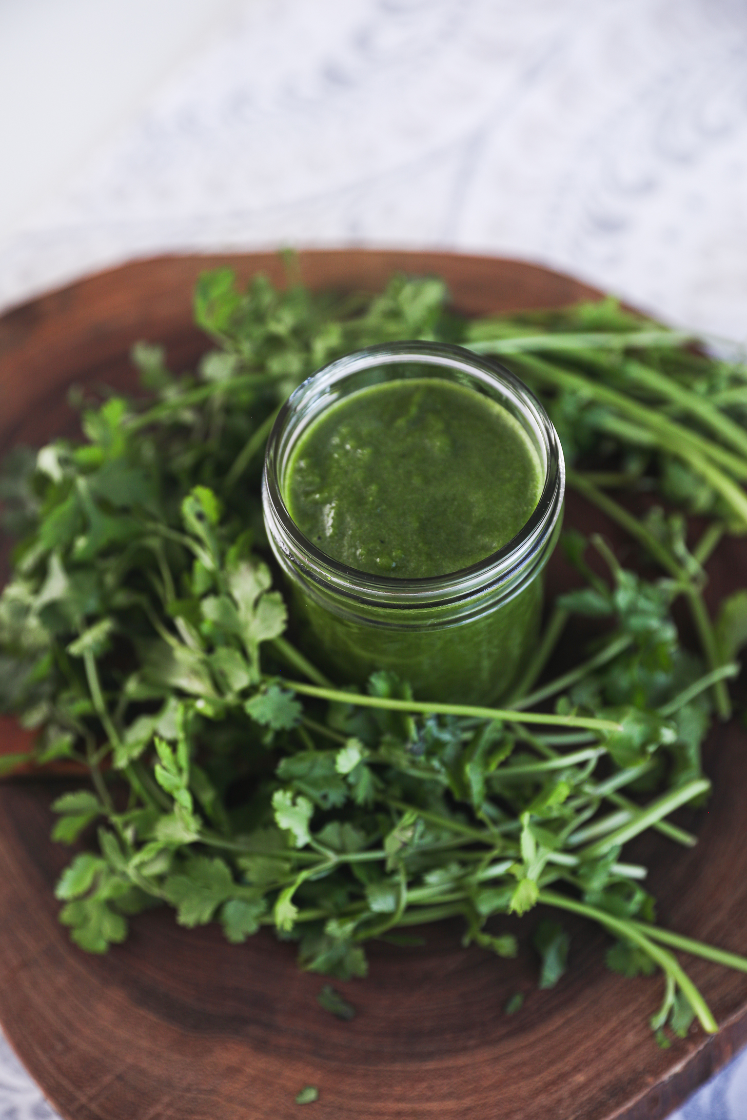 A mason jar of green chutney (sauce) surrounded by fresh cilantro on a wooden board.