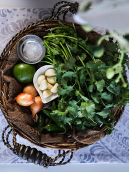 Top view image of a basket of food ingredients including fresh cilantro, garlic, shallots, lime, chillies and salt.