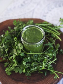 Perspective image of a jar of green chutney encircled by frehs cilantro on top of a wooden baord.