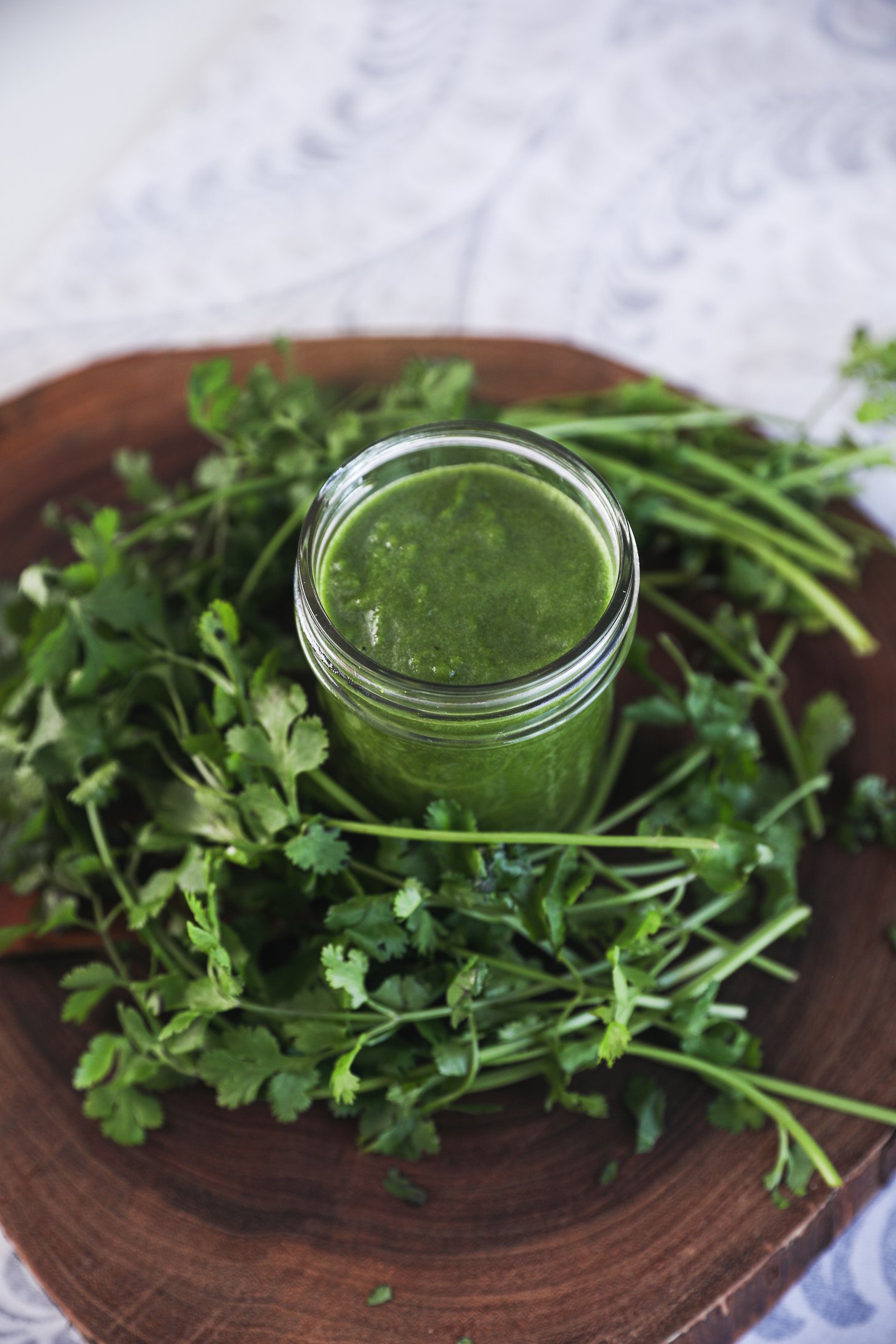 Perspective image of a jar of green chutney encircled by frehs cilantro on top of a wooden baord.