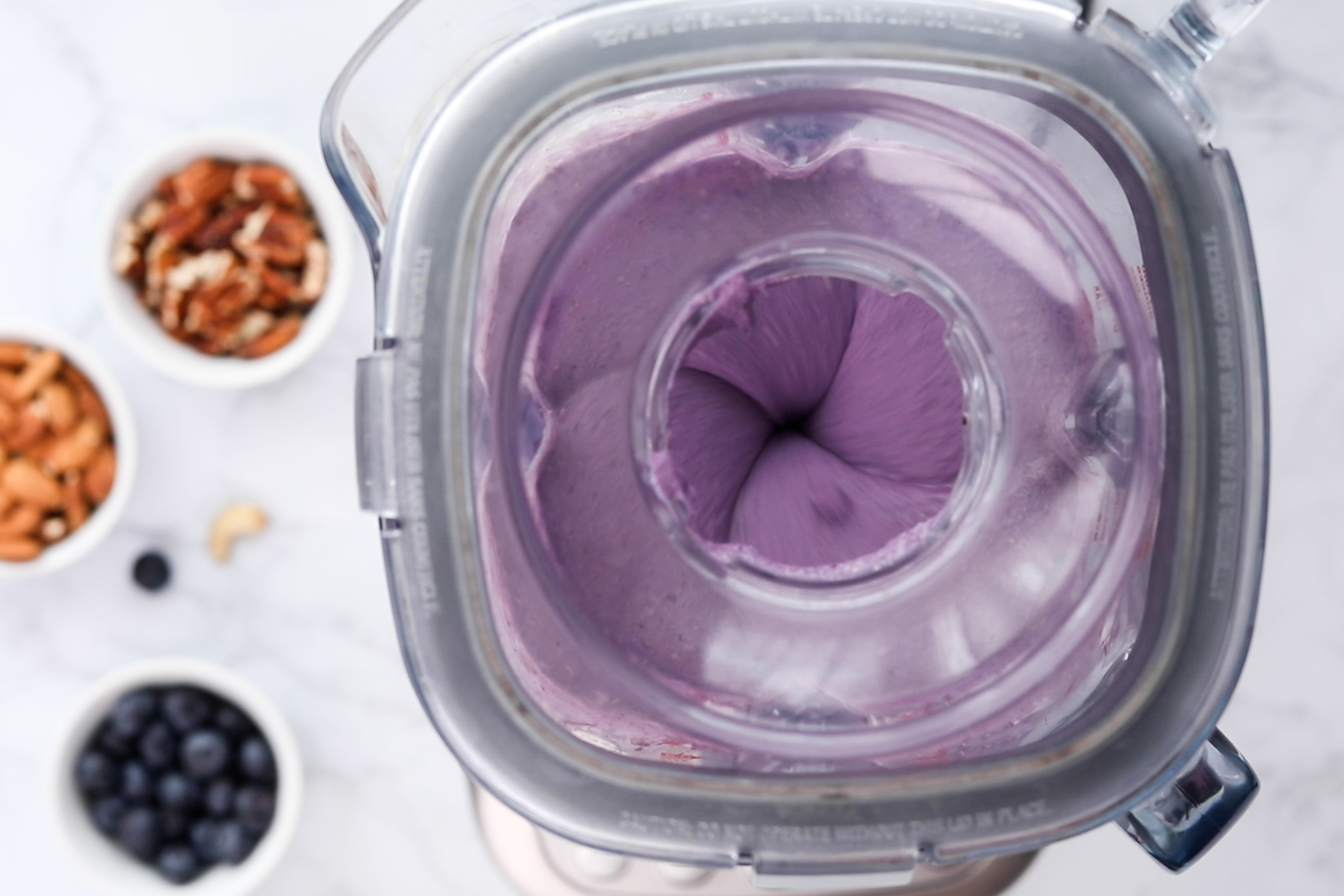 Top-down image capturing a blender in action blending creamy blueberry filling, with ramekins of nuts and berries visible in the background.