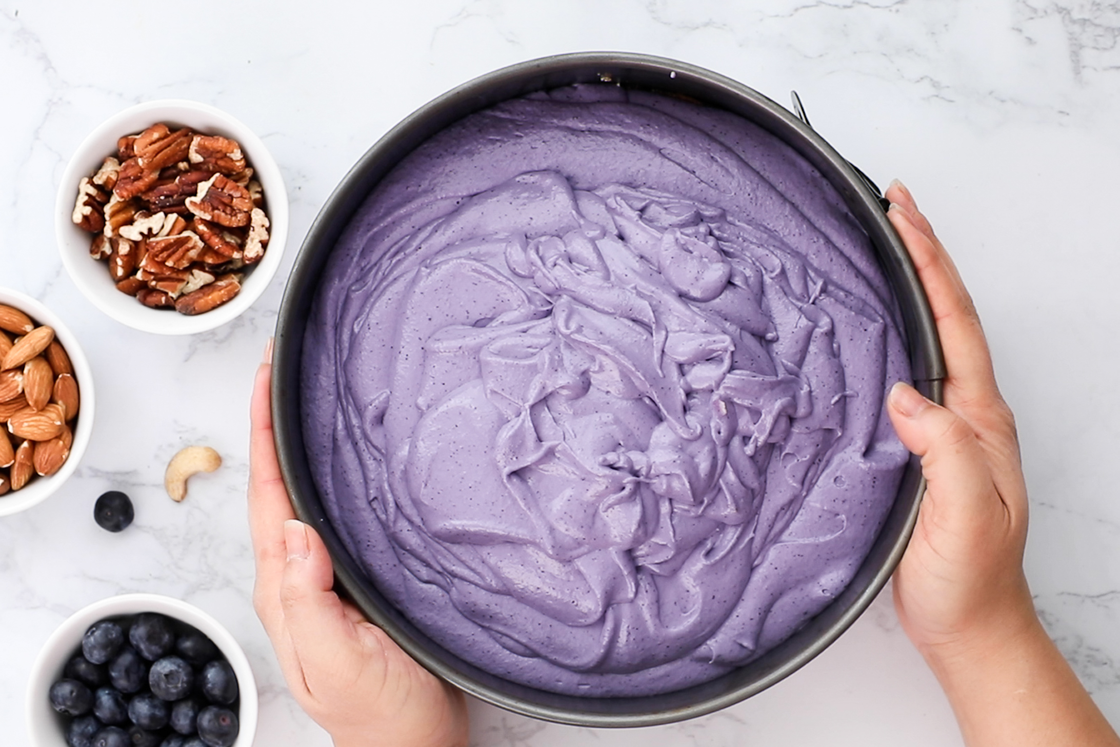Top-down image featuring hands holding a cake pan filled with creamy blueberry filling, accompanied by ramekins of nuts and berries nearby.