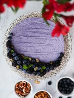 Top-down image of a blueberry cheesecake on a gold plate. One side is topped with berries. Nuts, berries, and flowers are in the foreground and background.
