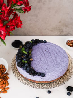 Perspective image of a blueberry cheesecake on a gold cake stand. One side is topped with berries. Nuts, berries, and flowers are in the foreground and background.