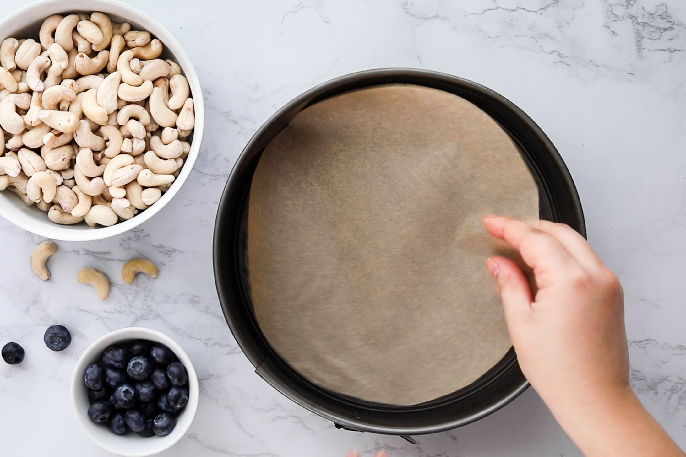 Hand placing a circular parchment paper into a cake pan with cashews and berries placed nearby.