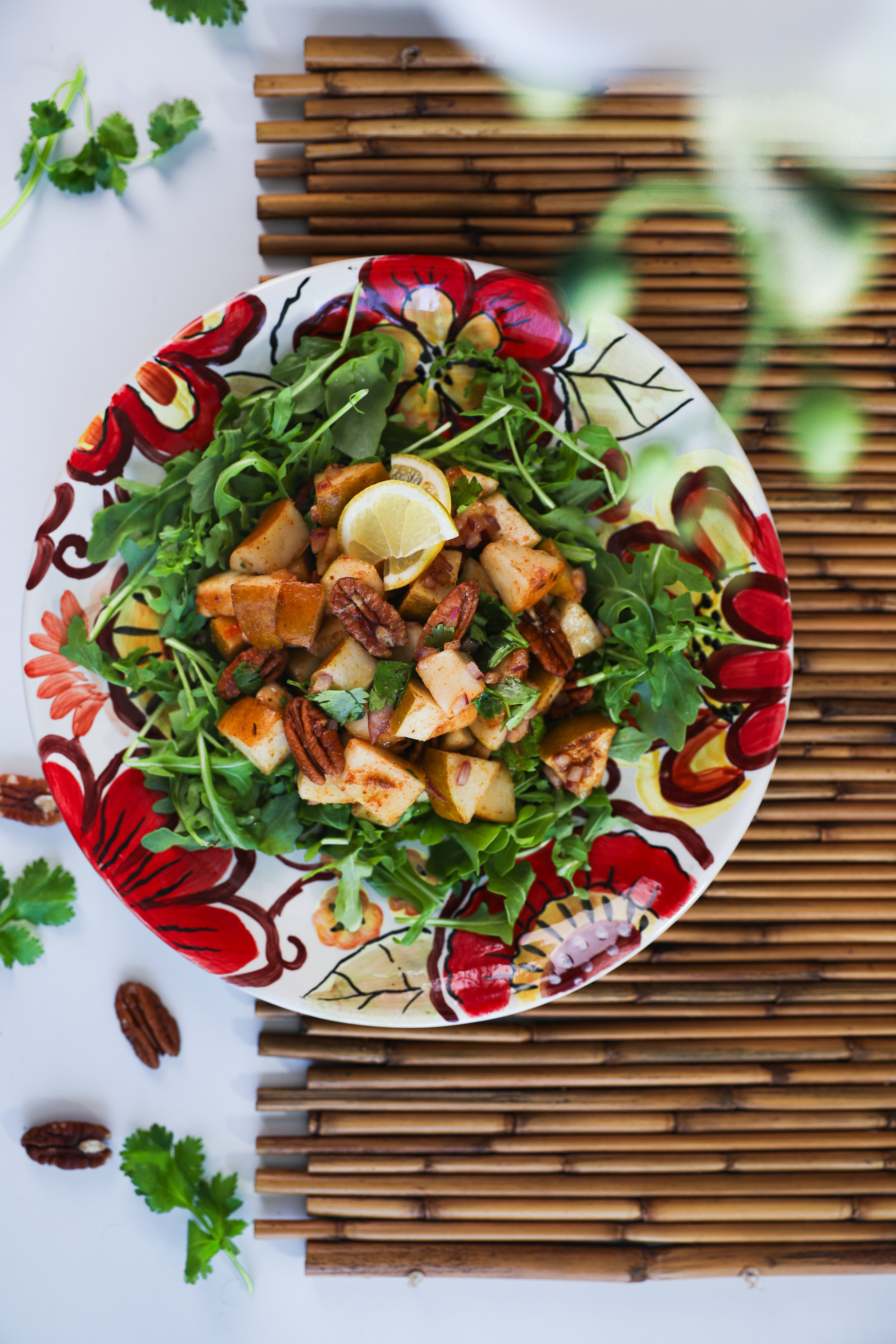 Pecan and pear salad arranged atop a bed of rocket (arugula), garnished with cilantro, pecans and lemon slices, served on a floral-printed plate.