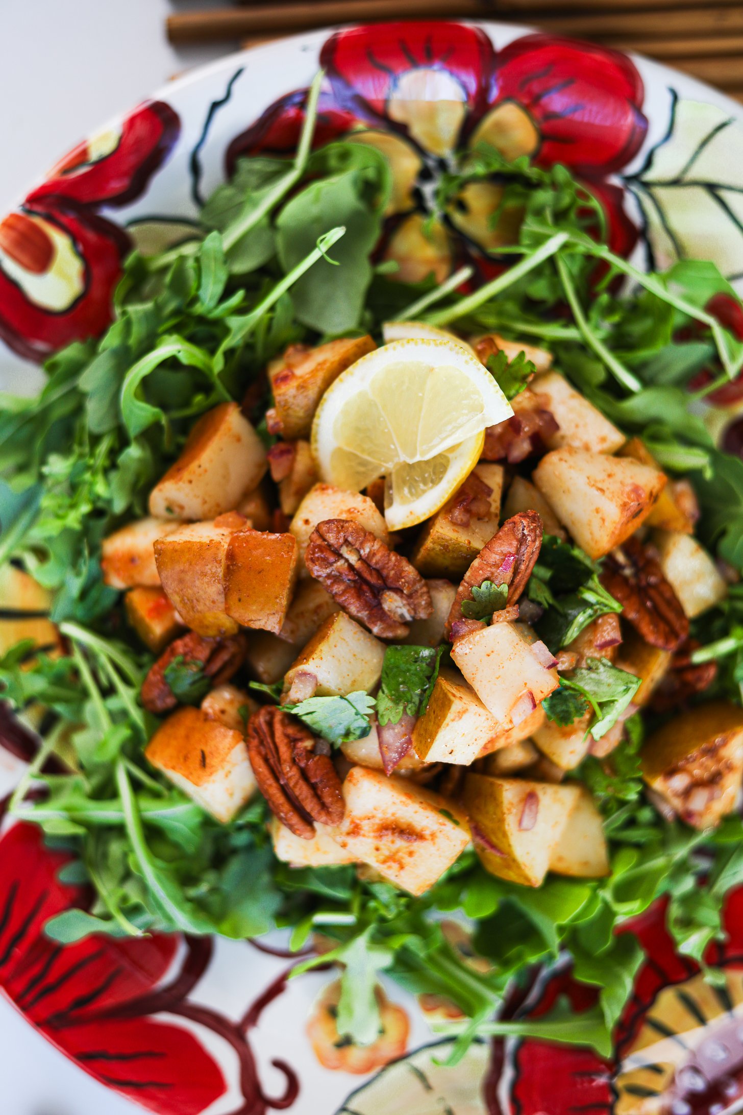 Top down view of Pecan and pear salad arranged atop a bed of rocket (arugula), garnished with cilantro, pecans and lemon slices, served on a floral-printed plate.