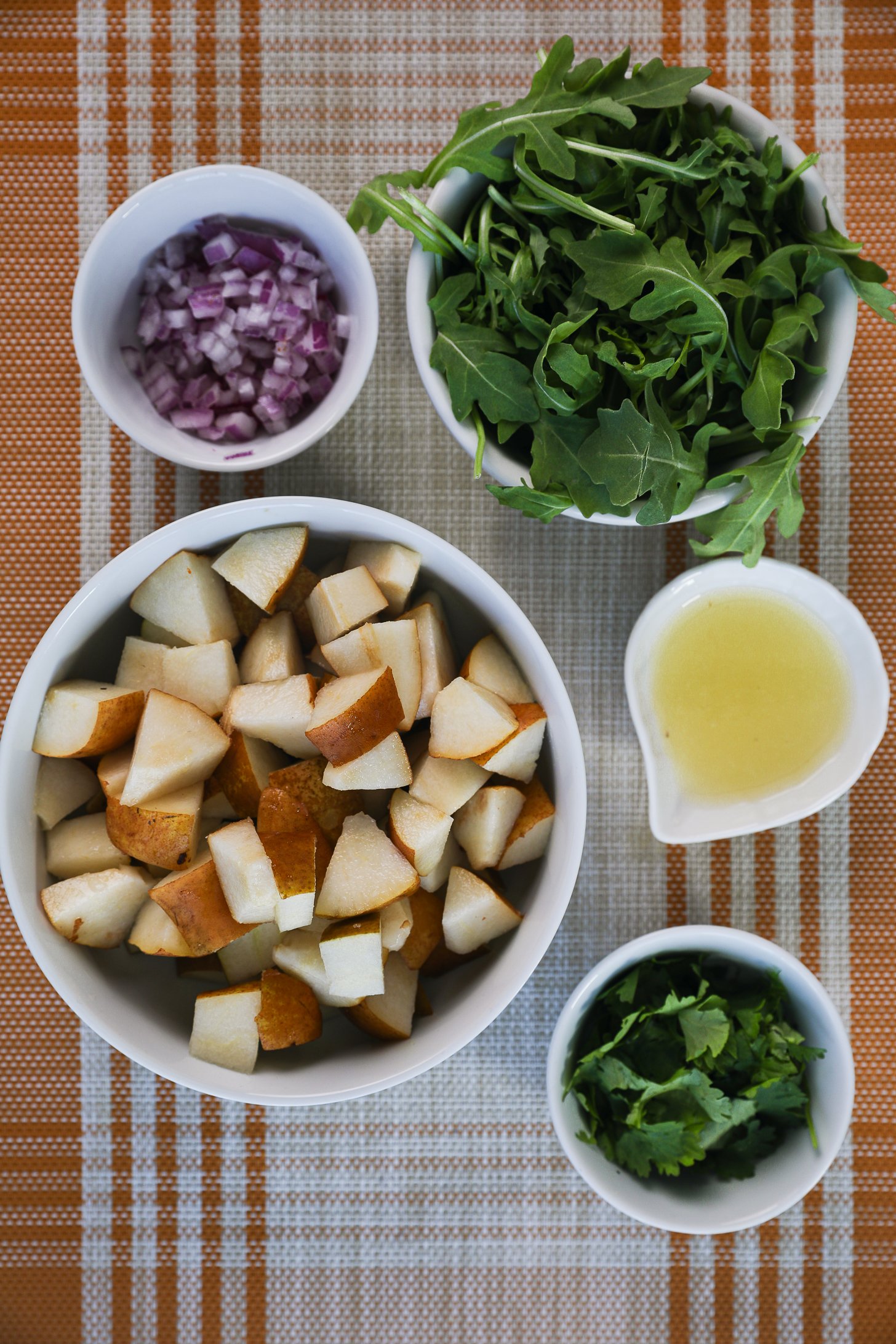 A display of food ingredients ready for salad prep. Ingredients include pear chunks, chopped red onion, rocket (arugula), lemon juice and cilantro leaves.