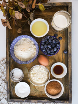 A selection of baking ingredients including flours, coconut sugar, oil, egg, milk and blueberries.
