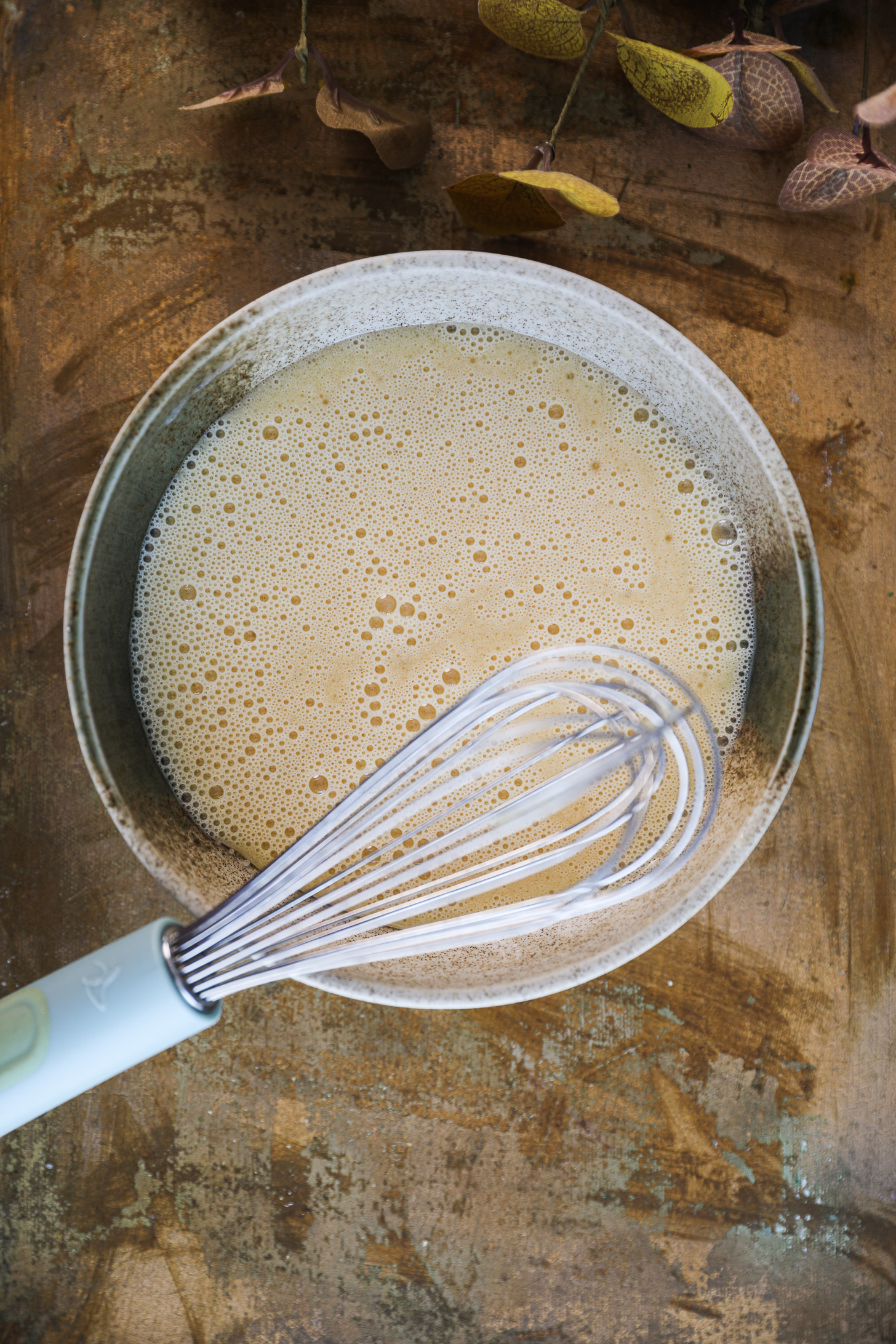 A bowl containing a frothy mixture of egg, milk, and oil, with a whisk resting on top.
