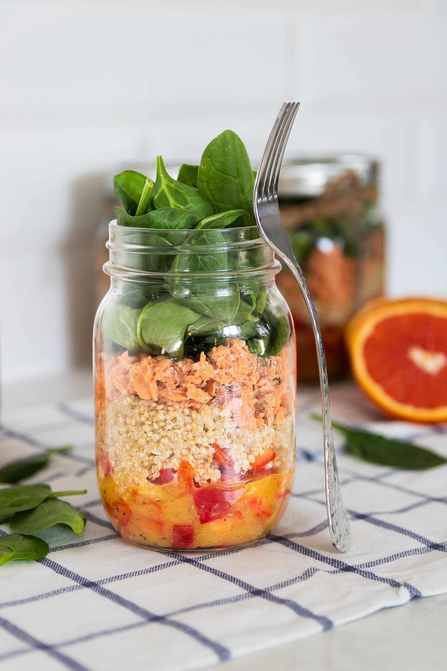A colourful layered mason jar salad with peppers, quinoa, canned salmon, and spinach leaves on top, with a fork leaning on the jar.