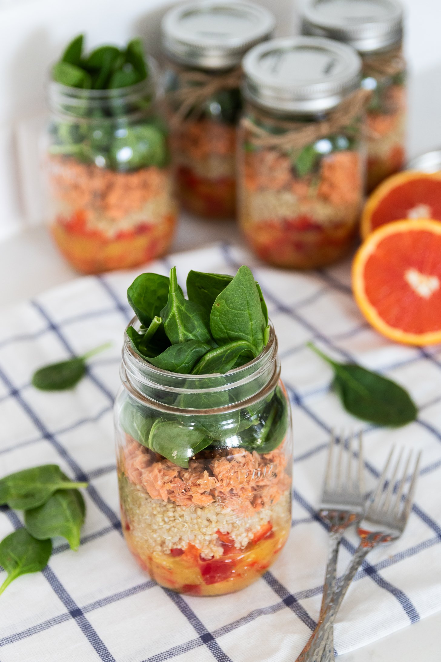A perspective image of five colourful layered mason jar salads with peppers, quinoa, canned salmon, and spinach leaves on top, creating a vibrant salad.
