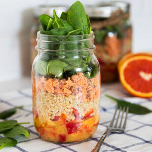 A colourful layered mason jar salad with peppers, quinoa, canned salmon, and spinach leaves on top, creating a vibrant salad.