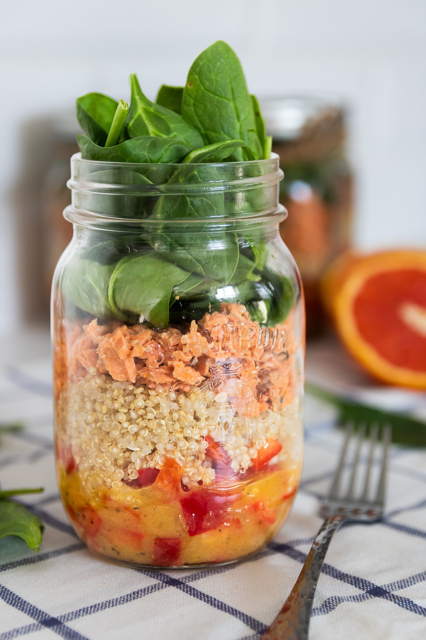 A close up image of a colourful layered mason jar salad with peppers, quinoa, canned salmon, and spinach leaves on top, creating a vibrant salad.