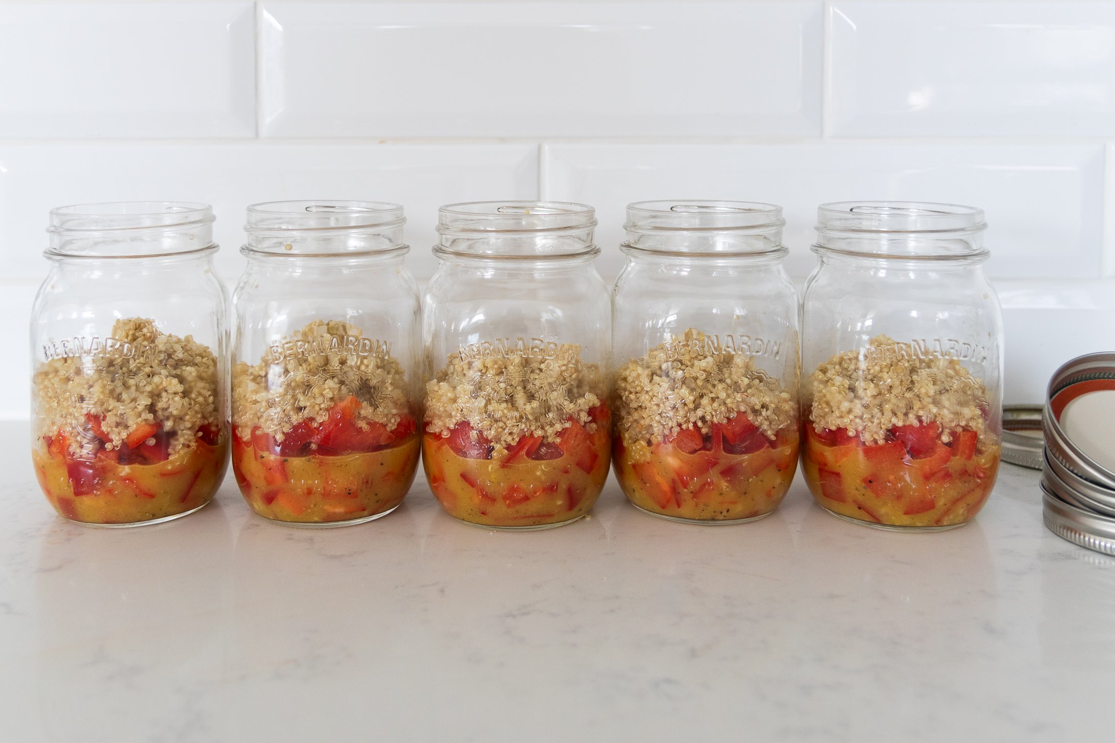 Five mason jars filled with dressing, peppers and quinoa leaning against a white background.