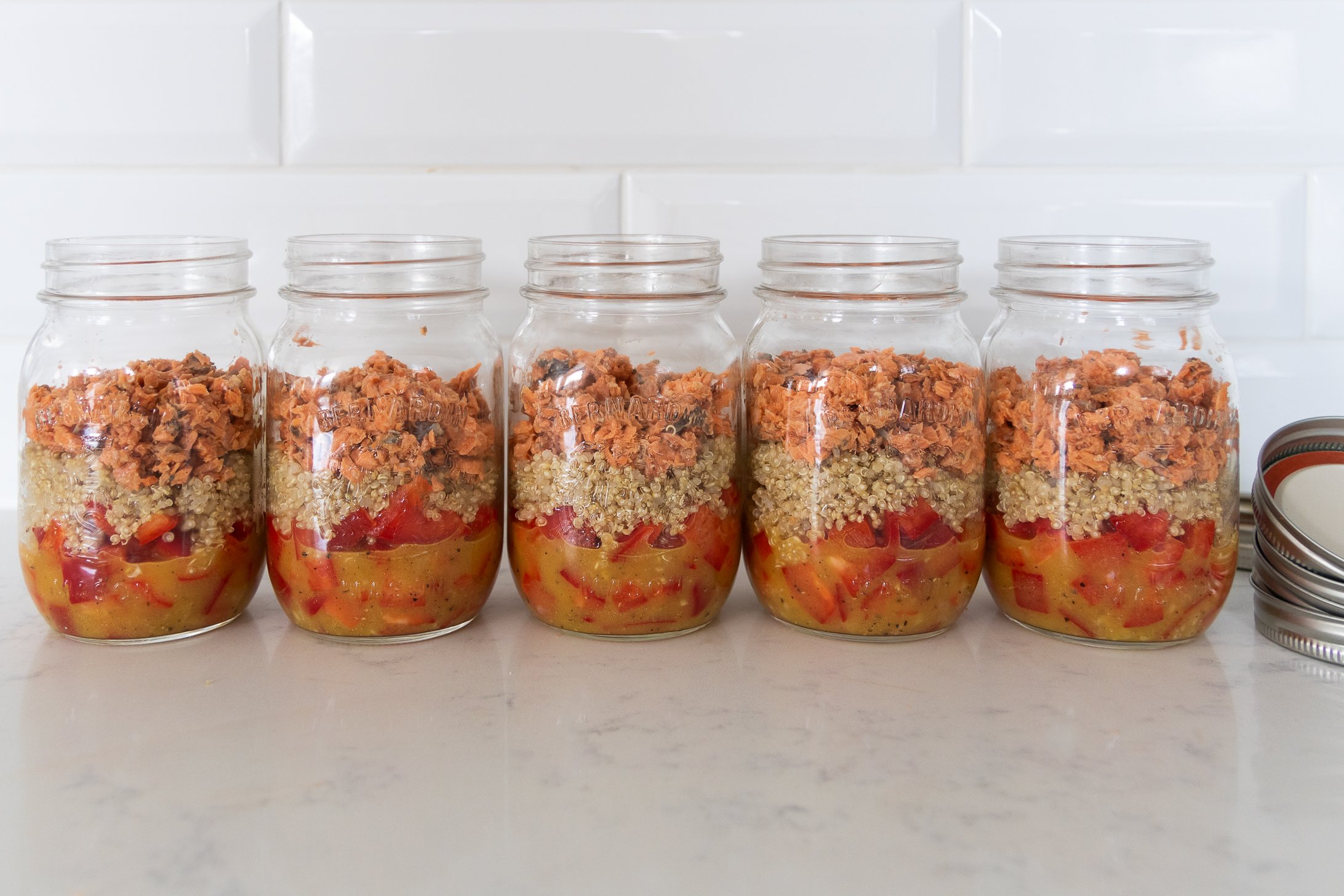 Five mason jars filled with dressing, peppers, quinoa and salmon leaning against a white background.