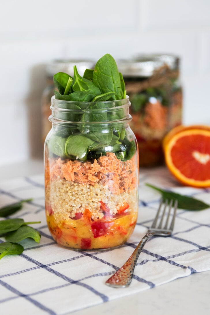 A colourful layered mason jar salad with peppers, quinoa, canned salmon, and spinach leaves on top, creating a vibrant salad.