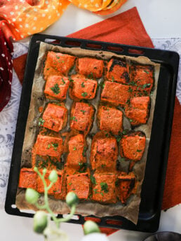 Overhead shot of a sheet pan featuring rows of salmon tandoori fish tikka, artfully styled on an orange mat with a traditional scarf.
