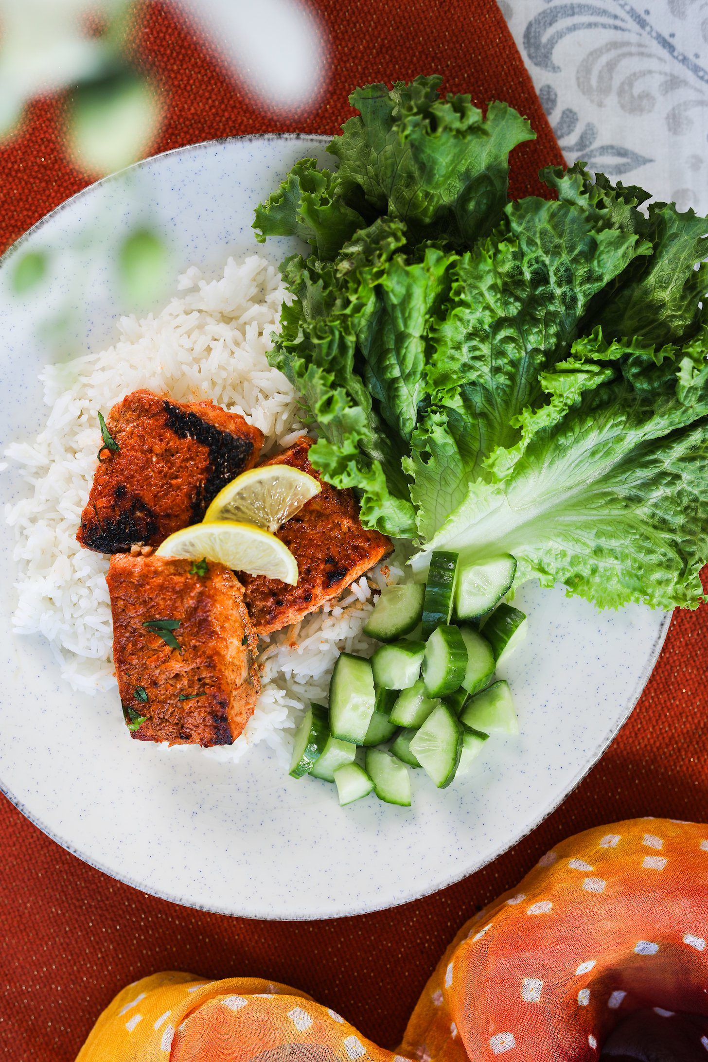 Overhead image of an arrangement of fish tikka pieces atop white rice, with a backdrop of lettuce leaves and cucumber chunks.