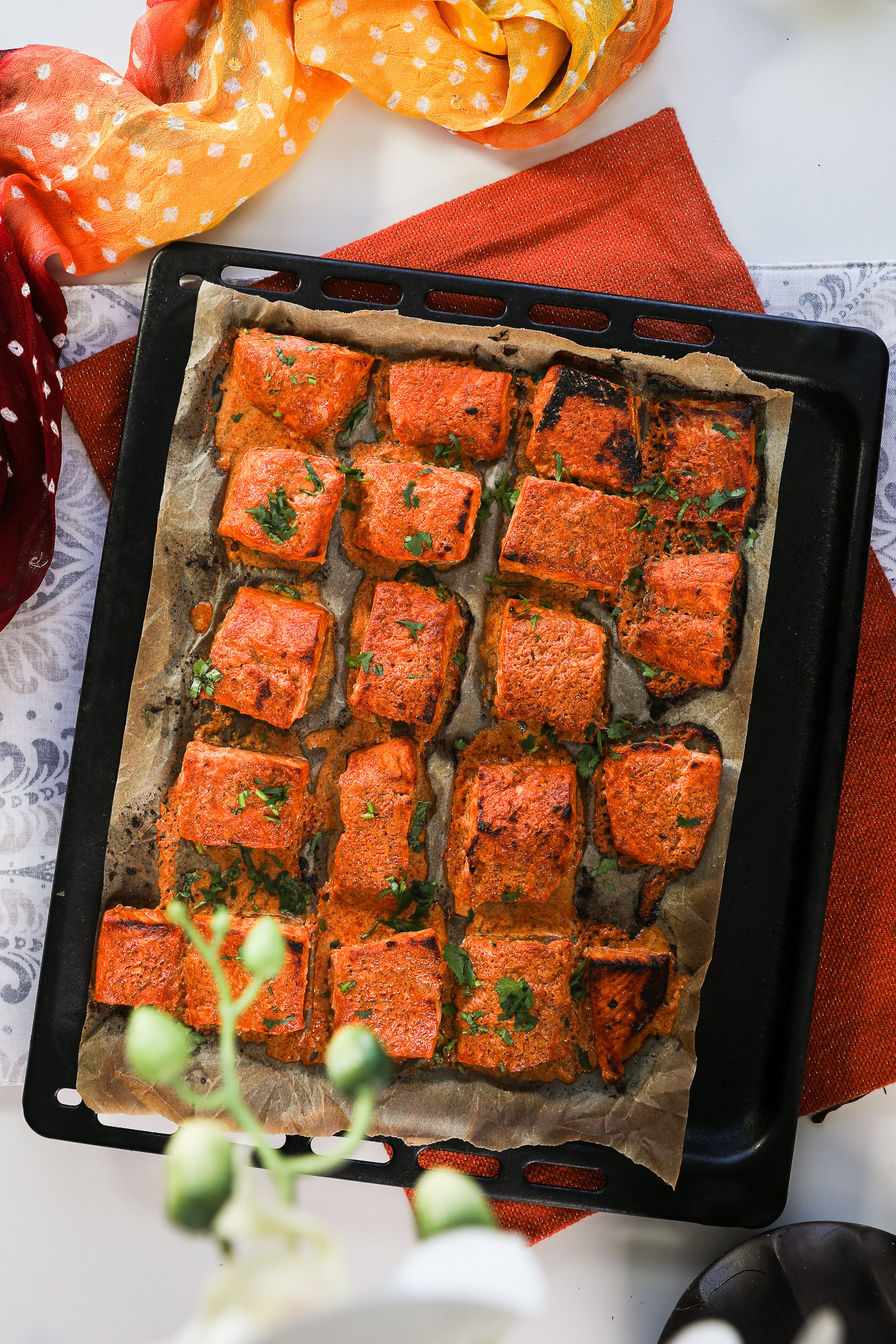 Overhead shot of a sheet pan featuring rows of salmon tandoori fish tikka, artfully styled on an orange mat with a traditional scarf.