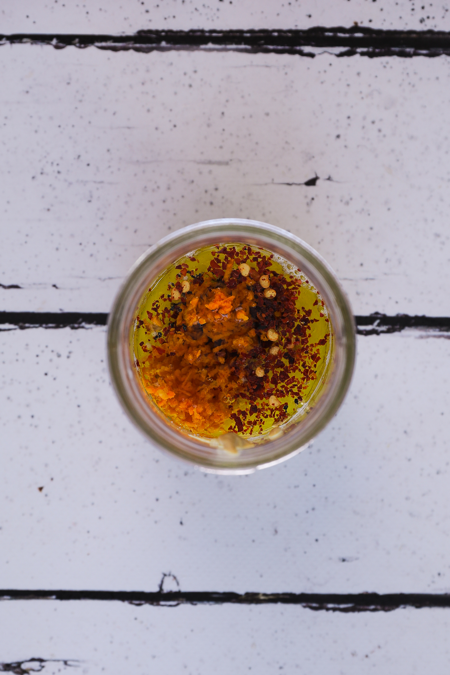 Top down view of an open mason jar showing lemon and oil topped with chilli flakes and grated fresh turmeric.