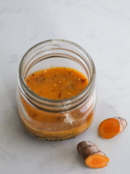 Perspective image of an open mason jar showing an orange-coloured dressing with fresh turmeric root nearby.