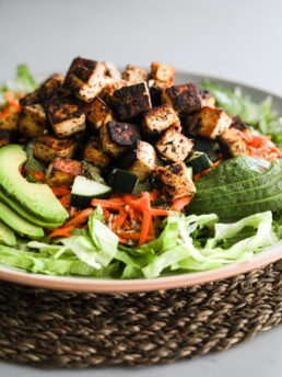 Perspective view of a generous salad plate featuring lettuce, avocado slices, carrot, and a pile of crispy tofu cubes on top.