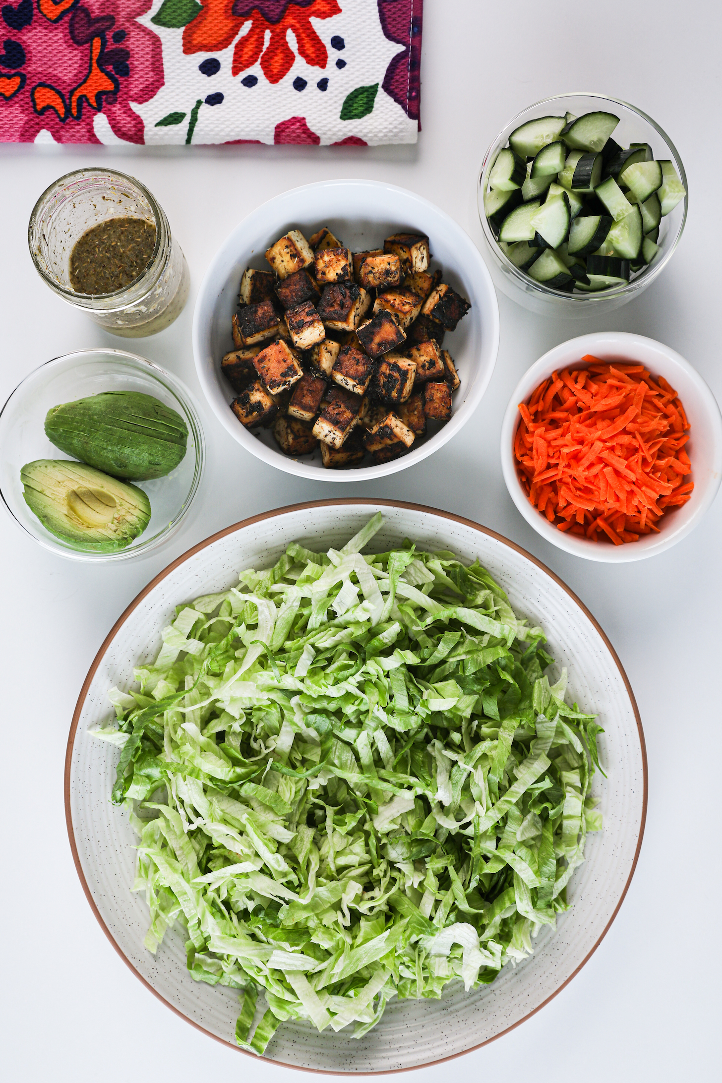A selection of ingredients featuring a large plate of shredded iceberg lettuce, grated carrot, crispy tofu cubes, cucumber cubes, avocado slices, and salad dressing.