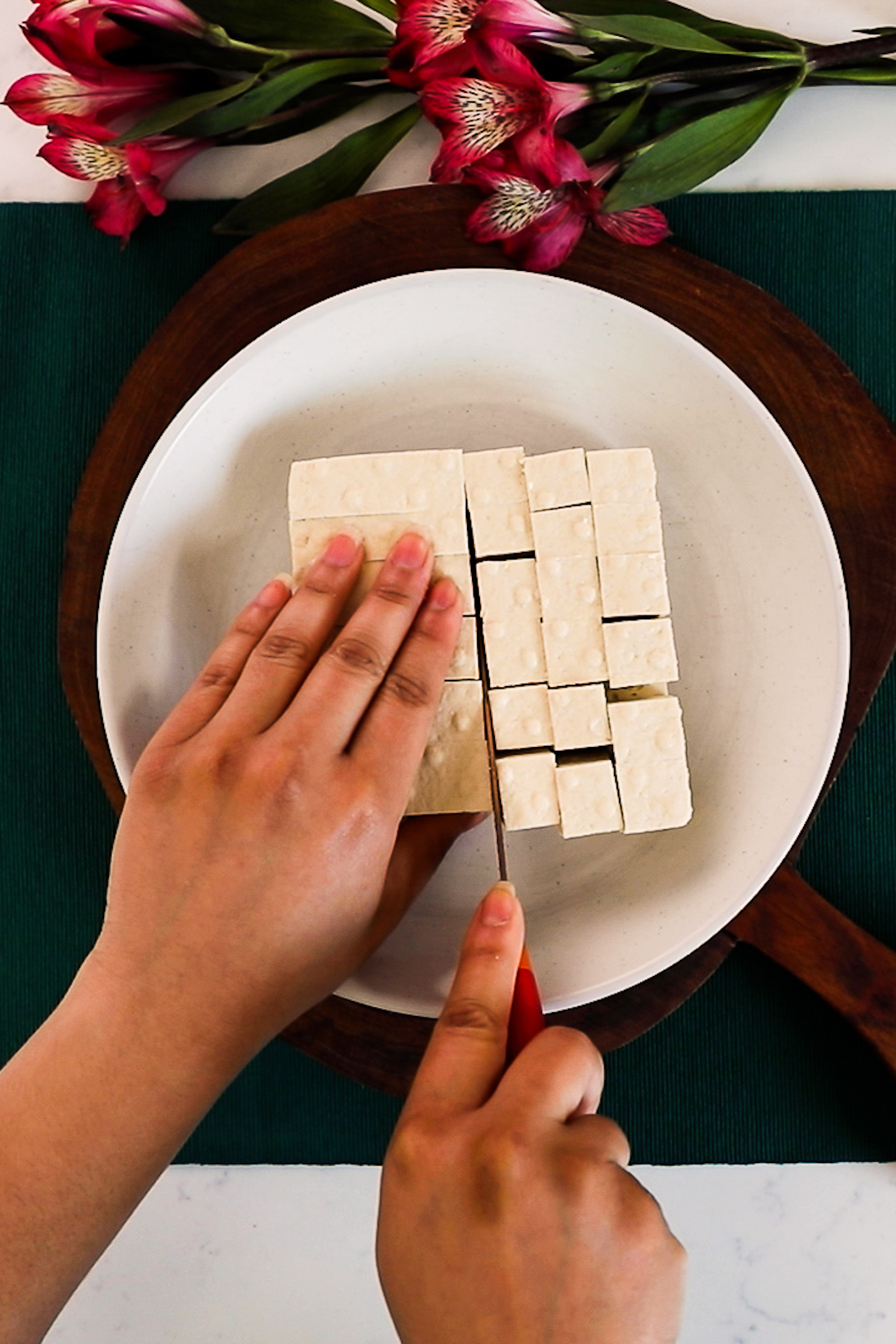 One hand holding a slab of tofu and the other cutting it into cubes using a knife.