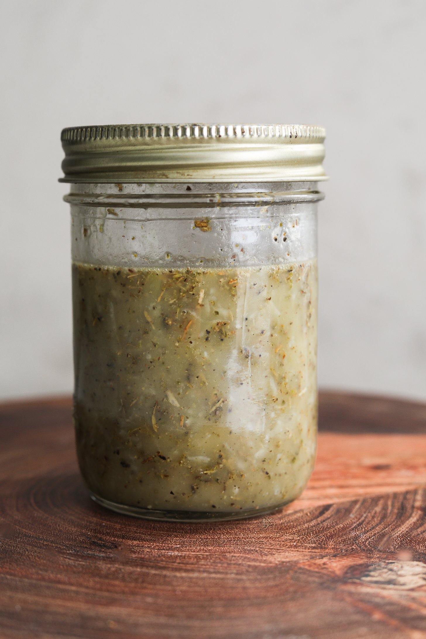 Close up image of a mason jar with lemon oil dressing containing floating dried herbs.