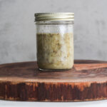 Perspective image of a mason jar with lemon oil dressing containing floating dried herbs.
