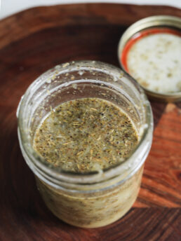Perspective image of an open mason jar filled with lemon oil dressing, with dried herbs floating inside.