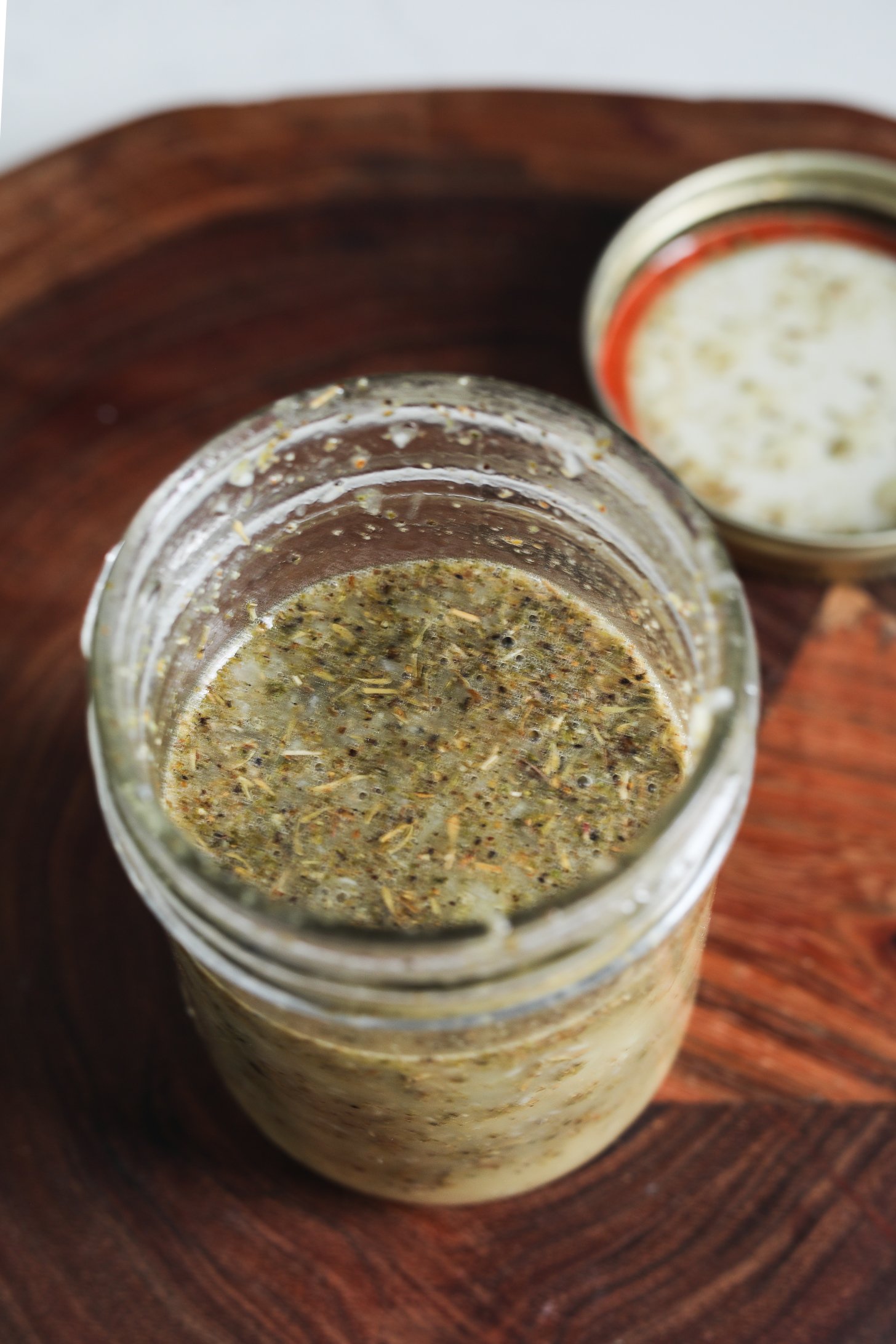 Perspective image of an open mason jar filled with lemon oil dressing, with dried herbs floating inside.