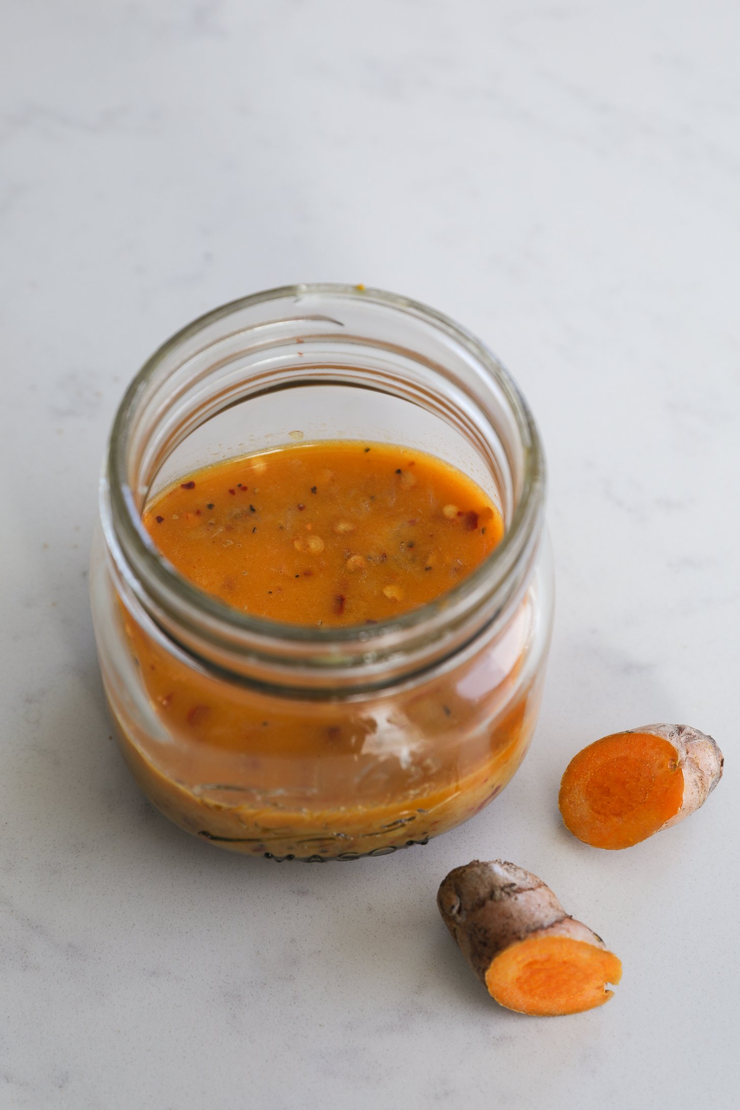A small mason jar filled with an orange coloured turmeric dressing with fresh turmeric root nearby.