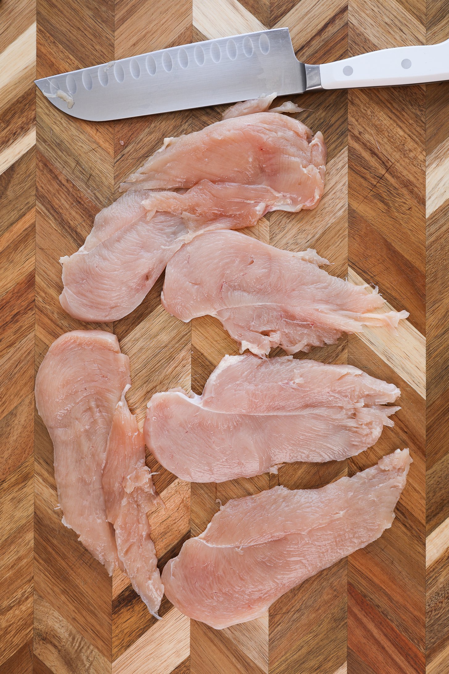 Thinly sliced chicken breasts laid on a wooden chopping board with a knife nearby.