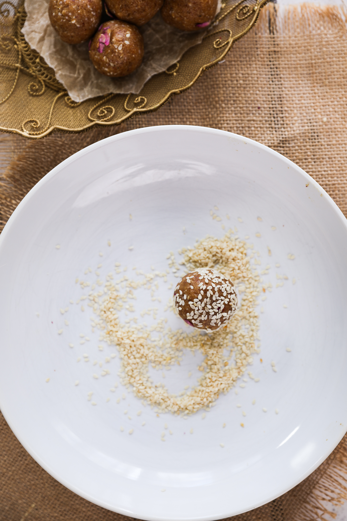 یک توپ بلیس قهوه‌ای پوشیده از کنجد که در میان دانه‌های کنجد روی یک بشقاب سفید قرار گرفته است.