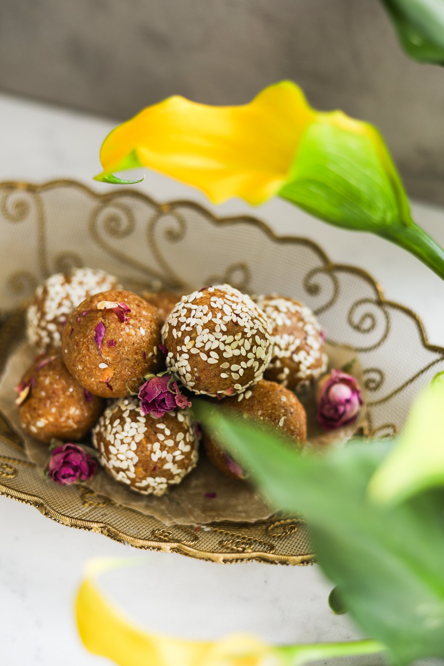 Gold oval tray with energy balls, half coated in sesame seeds, adorned with dried roses, beside yellow lilies.
