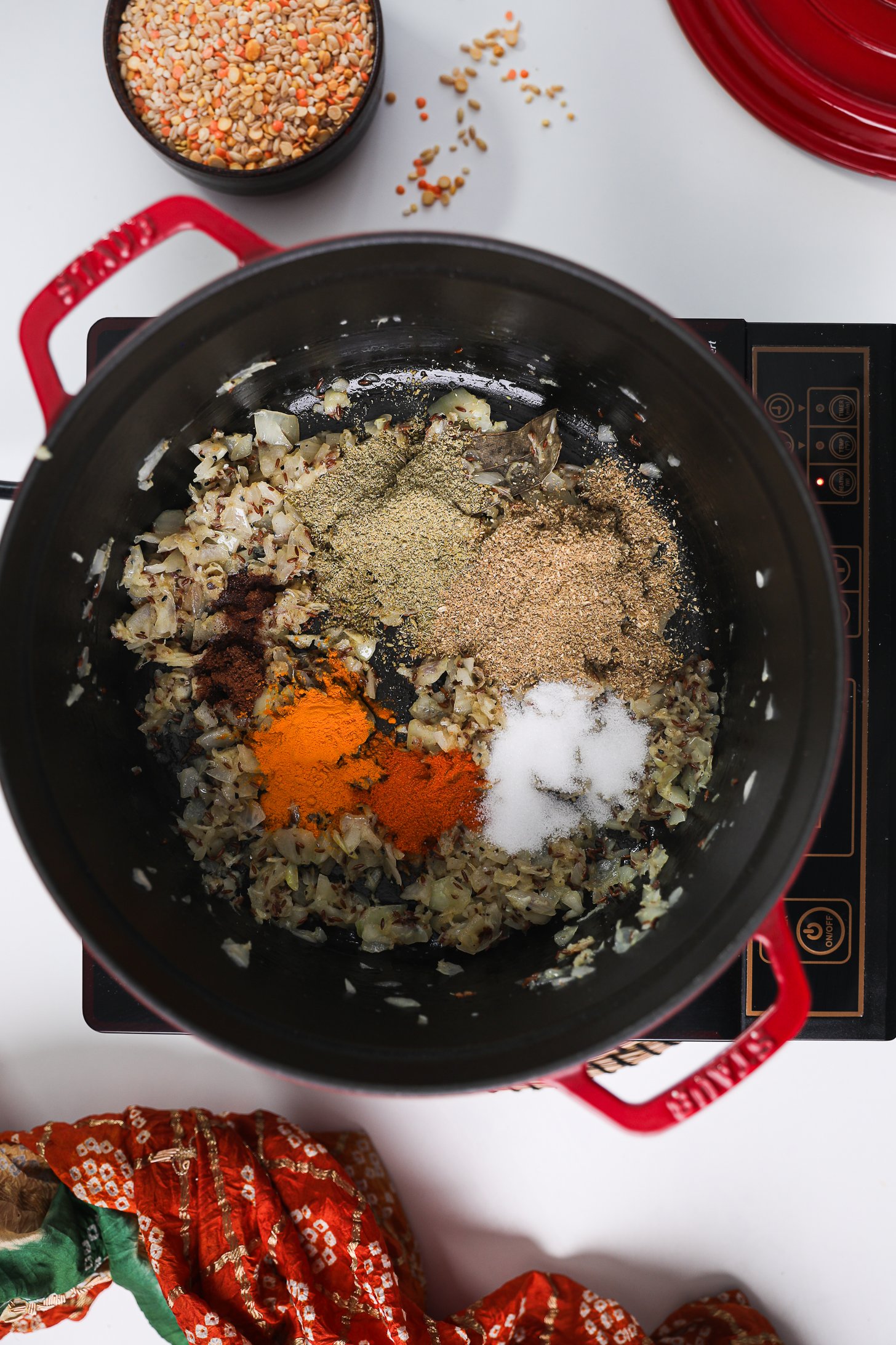 Indian powdered spices on top of fried onions and seeds in a cook pot on a mobile cooktop.