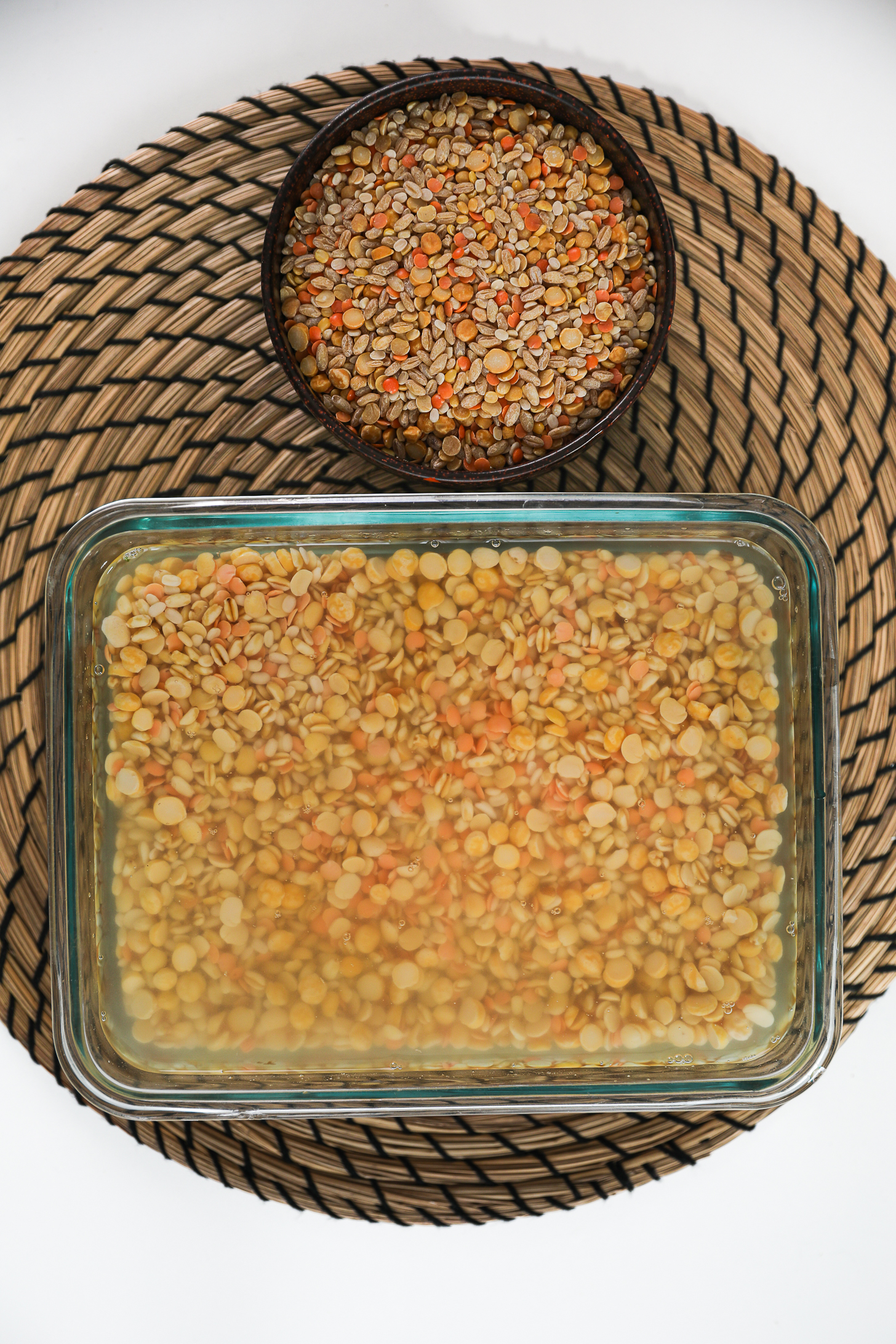 Two containers of lentils and wheat on a round straw mat: one soaked and the other dry.