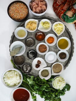 Indian spices on a round silver tray with bowls of chicken, lentils, onion, and tomato nearby.