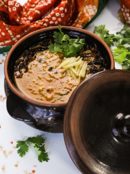 Perspective image of a serving vessel with haleem, topped with fried onions, ginger strips, and cilantro leaves.