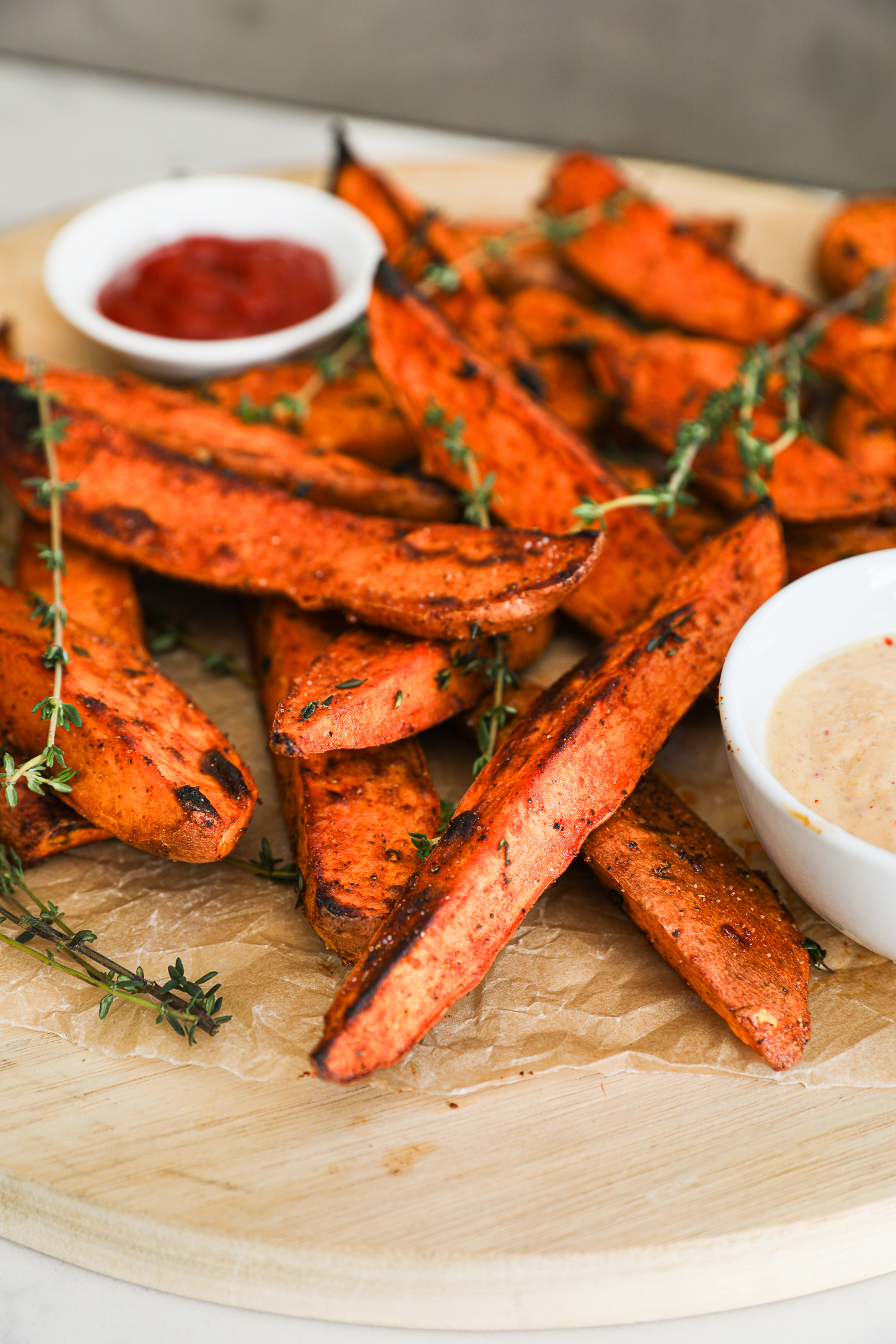 A close-up photo featuring golden-brown sweet potato wedges, garnished with fresh thyme sprigs, accompanied by two dipping sauces nearby.