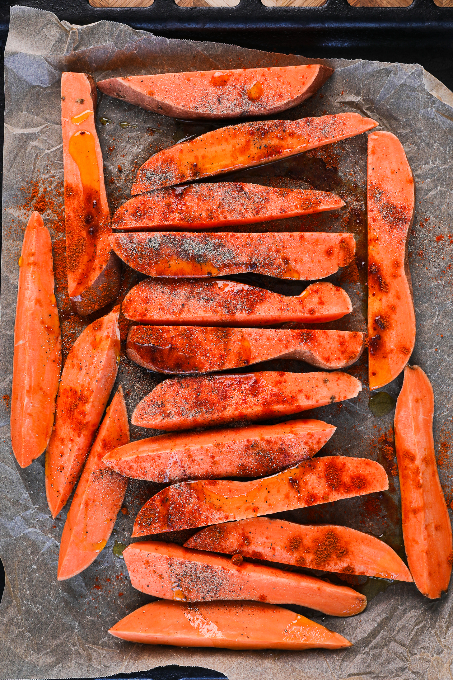 Uncooked sweet potato wedges with oil and powdered spices on top on a lined baking tray.