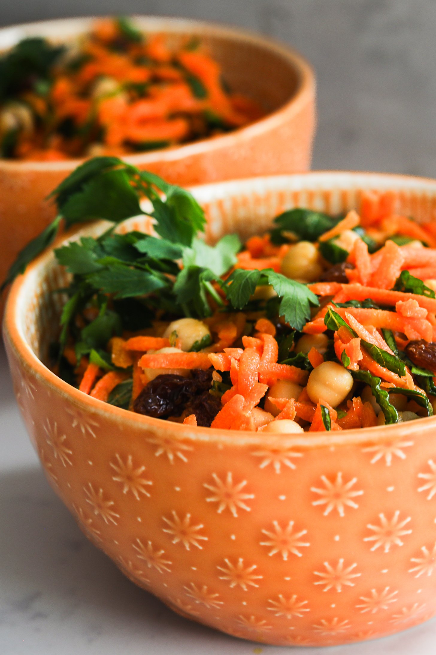 Close-up of two orange bowls filled with grated carrot salad with chickpeas, raisins, and herbs.