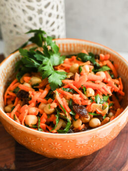 Angled close-up of a bowl of grated carrot salad with chickpeas, raisins, and herbs.