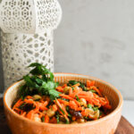 Perspective image of an orange bowl of grated carrot salad with raisins, chickpeas and herbs.